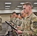 A group of U.S. Army soldiers in Operational Camouflage Pattern (OCP) uniforms are standing in rows inside an indoor facility, reading from black-framed documents or certificates. The setting appears to be a formal military ceremony, likely a promotion, award, or oath-of-office event. Multiple soldiers of varying ranks and backgrounds are visible, all focused on the documents they are holding.