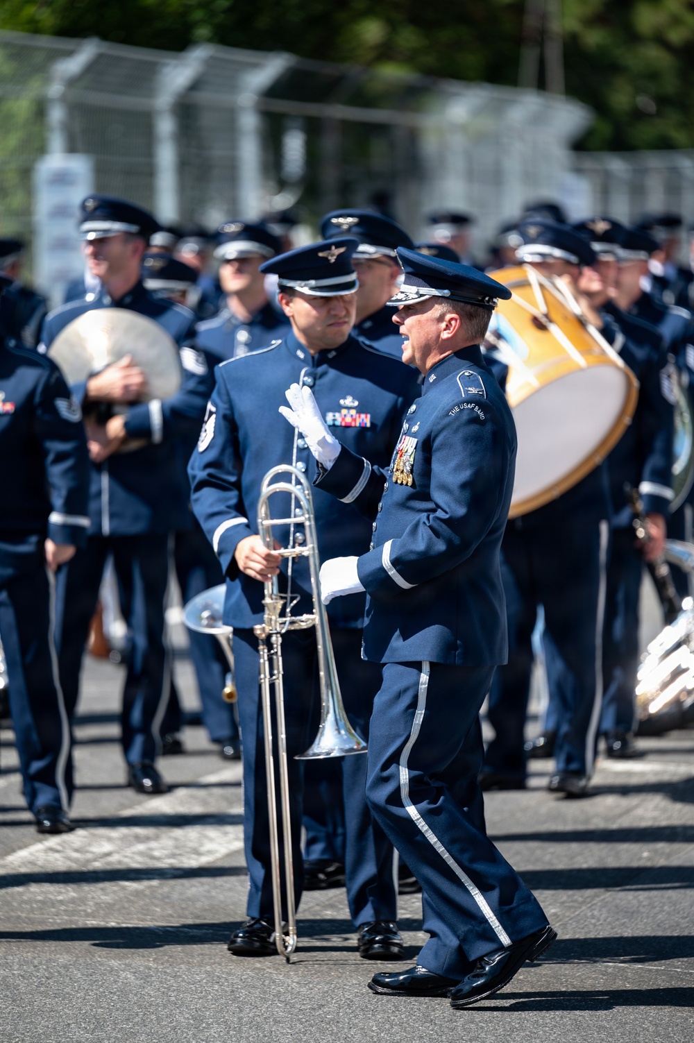 A United States Air Force Band is marching in a parade or ceremonial procession on a paved road. The foreground features a senior officer in dress uniform with white gloves and multiple medals, appearing to interact with a trombone player. The band members are dressed in formal USAF blue dress uniforms with service caps.