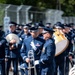 A United States Air Force Band is marching in a parade or ceremonial procession on a paved road. The foreground features a senior officer in dress uniform with white gloves and multiple medals, appearing to interact with a trombone player. The band members are dressed in formal USAF blue dress uniforms with service caps.
