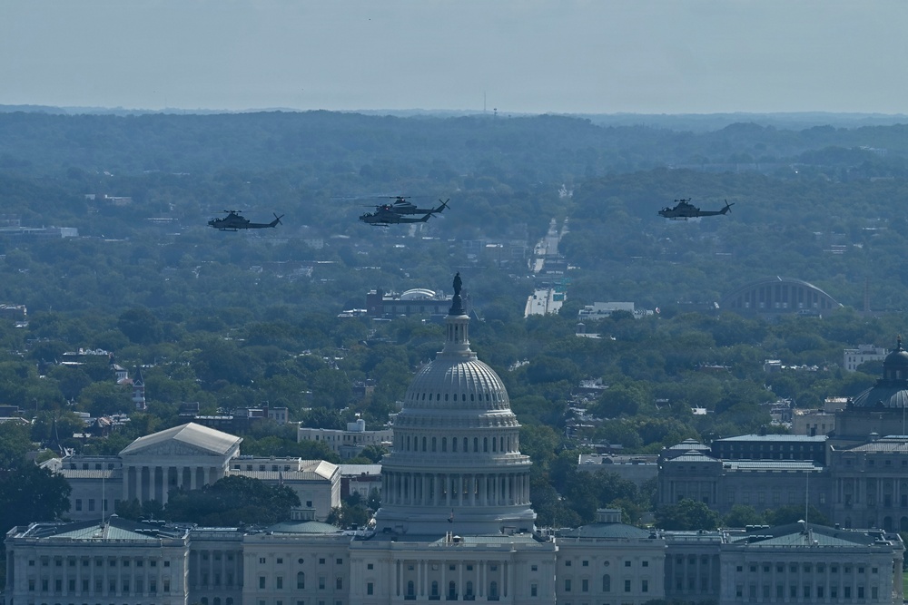 An aerial photograph shows three military helicopters flying in formation over Washington D.C., with the United States Capitol Building prominently visible in the foreground. The image appears to capture a flyover event, likely associated with a military ceremony or national celebration. The cityscape extends into the background with dense tree cover and urban infrastructure visible.