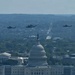 An aerial photograph shows three military helicopters flying in formation over Washington D.C., with the United States Capitol Building prominently visible in the foreground. The image appears to capture a flyover event, likely associated with a military ceremony or national celebration. The cityscape extends into the background with dense tree cover and urban infrastructure visible.