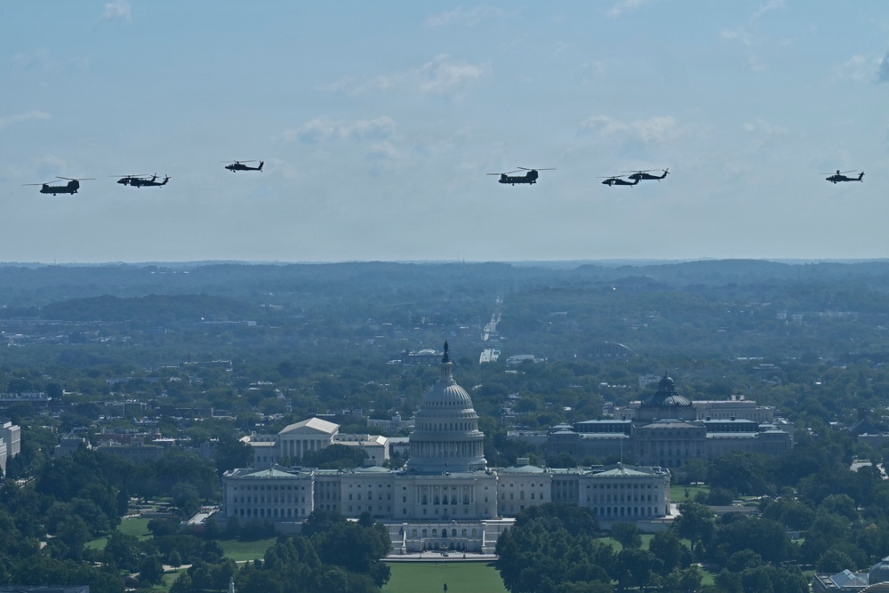 An aerial photograph shows a formation of military helicopters flying over Washington D.C., with the United States Capitol building prominently visible in the foreground. The helicopters appear to be conducting a flyover or parade formation, likely for a ceremonial or commemorative event. The image is taken from an elevated vantage point, possibly from the Washington Monument or a nearby tall structure.