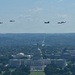 An aerial photograph shows a formation of military helicopters flying over Washington D.C., with the United States Capitol building prominently visible in the foreground. The helicopters appear to be conducting a flyover or parade formation, likely for a ceremonial or commemorative event. The image is taken from an elevated vantage point, possibly from the Washington Monument or a nearby tall structure.