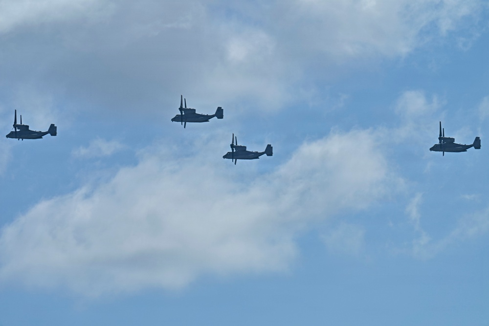 Four military tiltrotor aircraft, identifiable as V-22 Osprey variants, are flying in a loose formation against a partly cloudy blue sky. The aircraft are captured in silhouette, showing their distinctive twin-rotor, fixed-wing configuration. This appears to be a military flight operation or formation exercise.