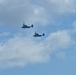 Four military tiltrotor aircraft, identifiable as V-22 Osprey variants, are flying in a loose formation against a partly cloudy blue sky. The aircraft are captured in silhouette, showing their distinctive twin-rotor, fixed-wing configuration. This appears to be a military flight operation or formation exercise.