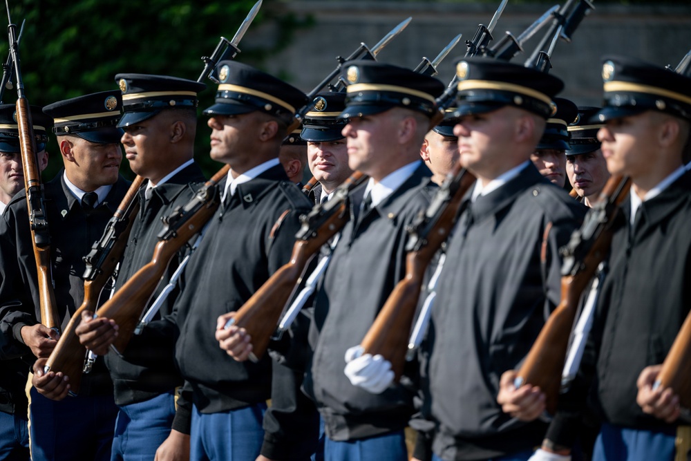 A formation of U.S. Army soldiers in ceremonial dress uniforms stands at attention during a formal military ceremony. The soldiers are equipped with rifles fitted with bayonets, held in a uniform carry position. The image captures a disciplined, organized military honor guard or ceremonial unit.