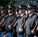 A formation of U.S. Army soldiers in ceremonial dress uniforms stands at attention during a formal military ceremony. The soldiers are equipped with rifles fitted with bayonets, held in a uniform carry position. The image captures a disciplined, organized military honor guard or ceremonial unit.