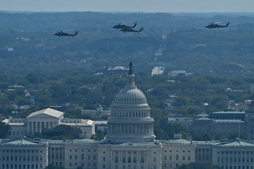 An aerial photograph shows three military helicopters flying in formation over Washington D.C., with the United States Capitol Building prominently visible in the foreground. The image appears to capture a military flyover event, likely during a national ceremony or parade. The Supreme Court building is also visible to the left of the Capitol.