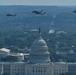 An aerial photograph shows three military helicopters flying in formation over Washington D.C., with the United States Capitol Building prominently visible in the foreground. The image appears to capture a military flyover event, likely during a national ceremony or parade. The Supreme Court building is also visible to the left of the Capitol.