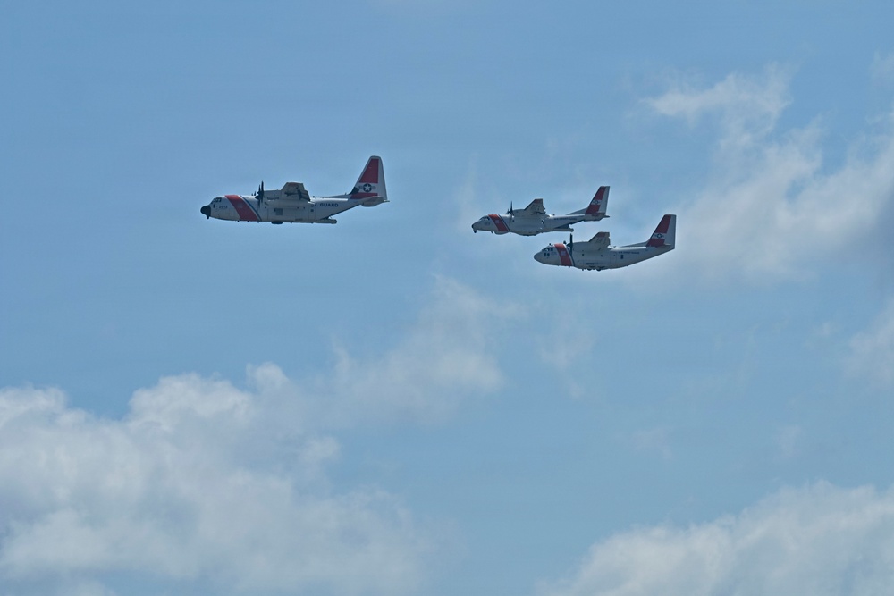 Three U.S. Coast Guard aircraft are flying in formation against a partly cloudy blue sky. The formation consists of one larger turboprop aircraft on the left, likely a C-130 Hercules, and two smaller aircraft flying in close proximity on the right. All aircraft display the distinctive white and red Coast Guard livery with the diagonal racing stripe markings.