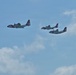 Three U.S. Coast Guard aircraft are flying in formation against a partly cloudy blue sky. The formation consists of one larger turboprop aircraft on the left, likely a C-130 Hercules, and two smaller aircraft flying in close proximity on the right. All aircraft display the distinctive white and red Coast Guard livery with the diagonal racing stripe markings.