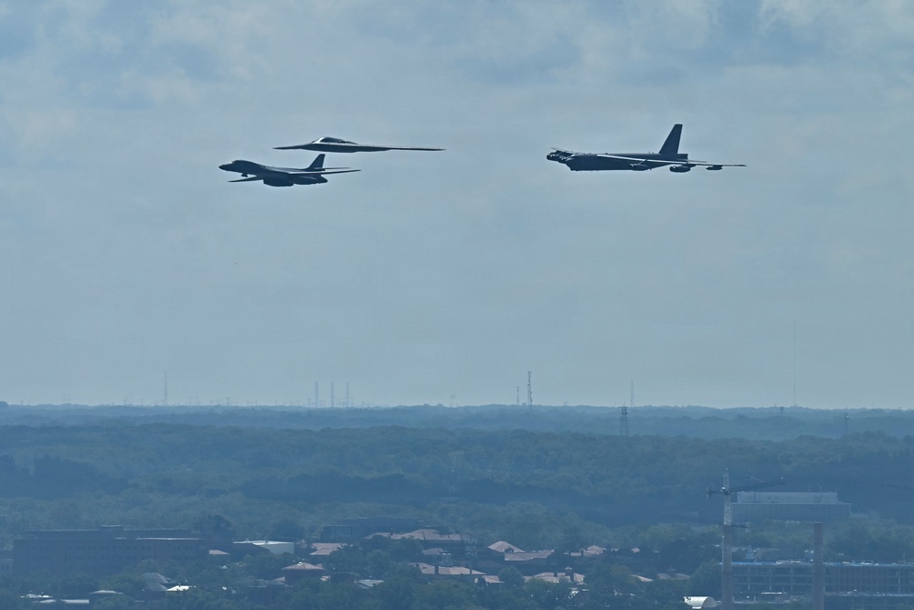 Three distinct US Air Force strategic bomber aircraft are flying in formation over a suburban landscape. The formation appears to include all three of America's current strategic bomber types flying together, a formation commonly known as a 'Bomb Trio' or 'Triad' flyover. The aircraft are silhouetted against a partly cloudy sky above a tree-lined suburban area.