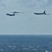 Three distinct US Air Force strategic bomber aircraft are flying in formation over a suburban landscape. The formation appears to include all three of America's current strategic bomber types flying together, a formation commonly known as a 'Bomb Trio' or 'Triad' flyover. The aircraft are silhouetted against a partly cloudy sky above a tree-lined suburban area.
