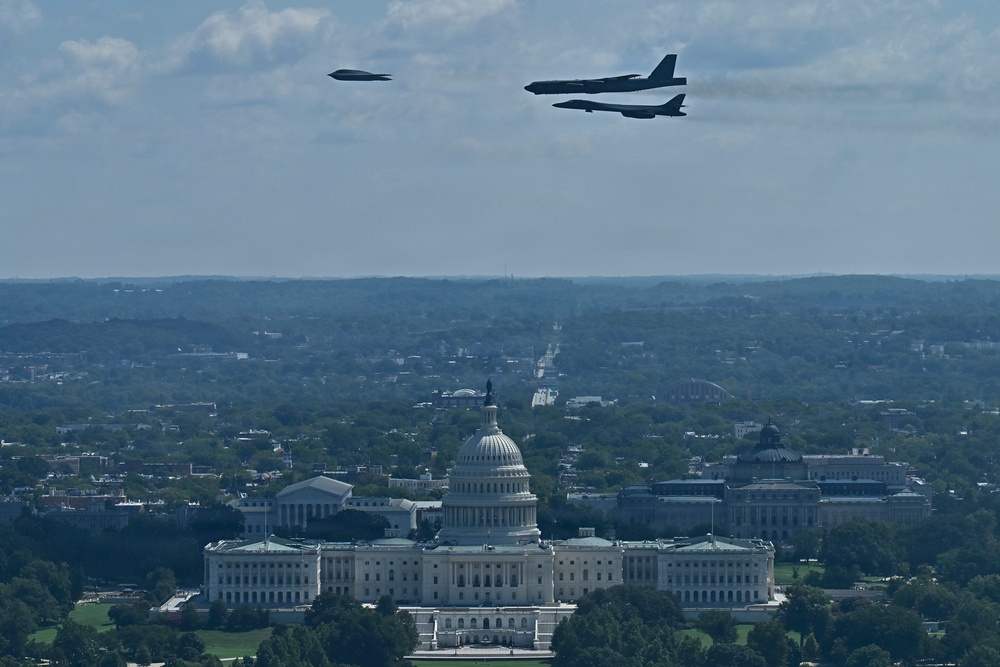 An aerial photograph shows military aircraft conducting a flyover above the United States Capitol building in Washington, D.C. Three aircraft are visible in formation — what appears to be a B-2 Spirit stealth bomber on the left and two B-52 Stratofortress bombers on the right. The image is taken from an elevated vantage point, likely the Washington Monument, providing a sweeping view of the National Mall and surrounding cityscape.