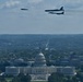 An aerial photograph shows military aircraft conducting a flyover above the United States Capitol building in Washington, D.C. Three aircraft are visible in formation — what appears to be a B-2 Spirit stealth bomber on the left and two B-52 Stratofortress bombers on the right. The image is taken from an elevated vantage point, likely the Washington Monument, providing a sweeping view of the National Mall and surrounding cityscape.