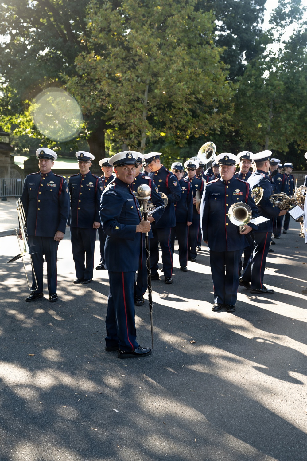 A military marching band is assembled outdoors on a paved path, likely in a park setting. The group is led by a drum major holding a ceremonial mace/baton staff at the front. Members are dressed in formal navy blue uniforms with red trim, gold buttons, and white peaked caps.