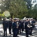 A military marching band is assembled outdoors on a paved path, likely in a park setting. The group is led by a drum major holding a ceremonial mace/baton staff at the front. Members are dressed in formal navy blue uniforms with red trim, gold buttons, and white peaked caps.