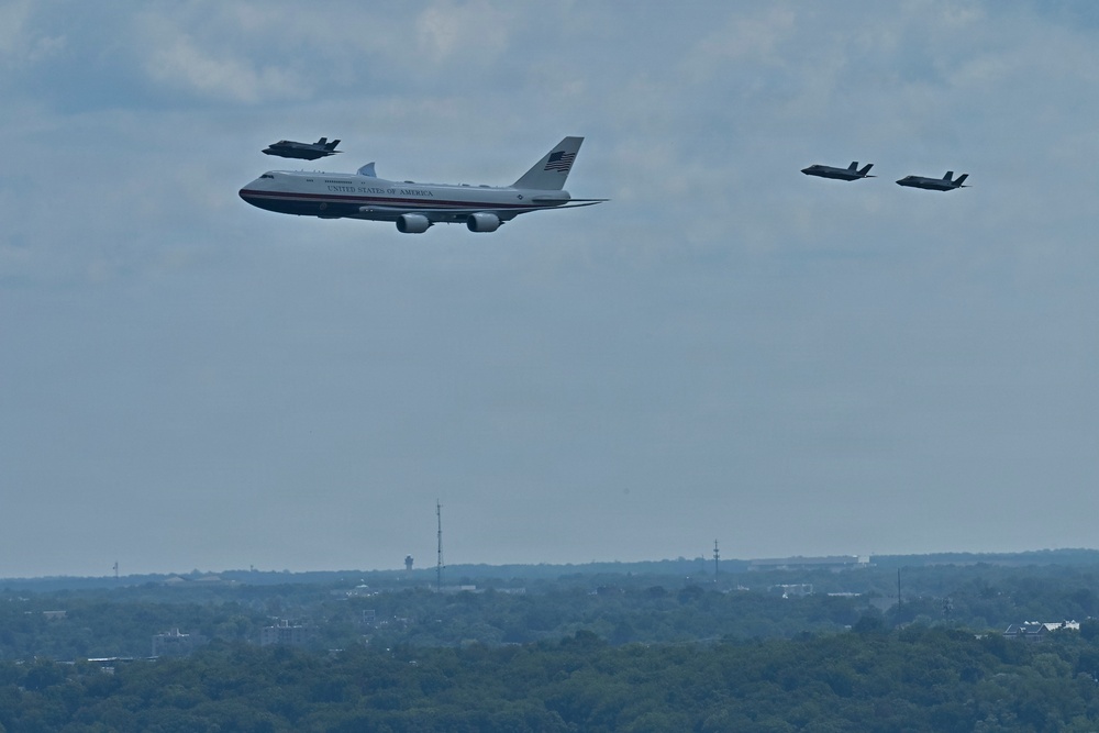 The image shows a large Boeing VC-25 aircraft marked 'United States of America' — commonly known as Air Force One — flying in formation with four F-35 fighter jets as escorts. The formation is photographed from a distance over a wooded suburban or urban landscape. The scene appears to depict an official flyover or transit flight.