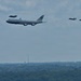 The image shows a large Boeing VC-25 aircraft marked 'United States of America' — commonly known as Air Force One — flying in formation with four F-35 fighter jets as escorts. The formation is photographed from a distance over a wooded suburban or urban landscape. The scene appears to depict an official flyover or transit flight.
