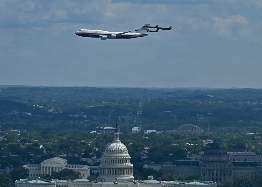 A large white aircraft marked 'United States of America' — consistent with Air Force One — flies over Washington D.C., with the U.S. Capitol dome prominently visible in the foreground. The image appears to be taken from an elevated vantage point, likely the Washington Monument observation deck. The cityscape, including the Supreme Court building to the left, stretches across the background beneath a partly cloudy sky.