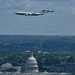 A large white aircraft marked 'United States of America' — consistent with Air Force One — flies over Washington D.C., with the U.S. Capitol dome prominently visible in the foreground. The image appears to be taken from an elevated vantage point, likely the Washington Monument observation deck. The cityscape, including the Supreme Court building to the left, stretches across the background beneath a partly cloudy sky.