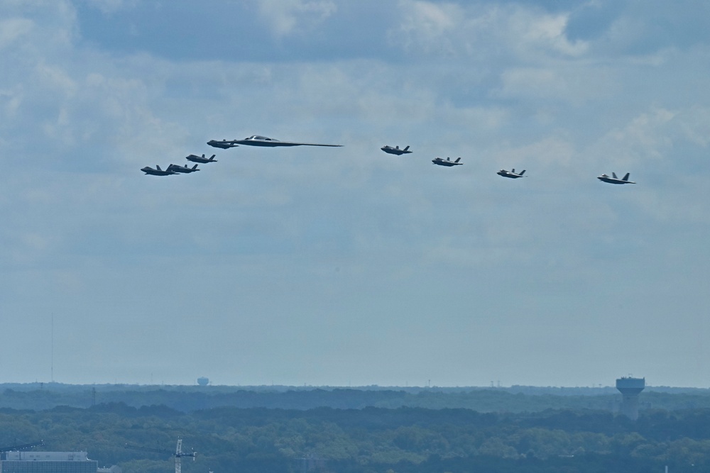 A formation of military aircraft is flying in a loose echelon or spread formation against a partly cloudy sky. The formation includes what appears to be a B-2 Spirit stealth bomber as the centerpiece, flanked by multiple smaller fighter jets, likely F-35s. The aircraft are flying at a moderate altitude over a flat, tree-lined landscape with urban infrastructure visible on the horizon.
