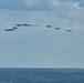 A formation of military aircraft is flying in a loose echelon or spread formation against a partly cloudy sky. The formation includes what appears to be a B-2 Spirit stealth bomber as the centerpiece, flanked by multiple smaller fighter jets, likely F-35s. The aircraft are flying at a moderate altitude over a flat, tree-lined landscape with urban infrastructure visible on the horizon.