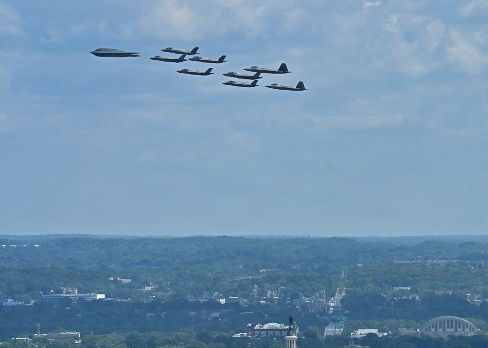 A formation of military aircraft flies over an urban landscape, likely Washington D.C., identifiable by visible monuments and architecture below. The formation appears to consist of a B-2 Spirit stealth bomber leading a group of F-35 and/or F-22 stealth fighter jets in a ceremonial flyover. The scene suggests a military parade or national celebration event.