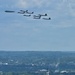 A formation of military aircraft flies over an urban landscape, likely Washington D.C., identifiable by visible monuments and architecture below. The formation appears to consist of a B-2 Spirit stealth bomber leading a group of F-35 and/or F-22 stealth fighter jets in a ceremonial flyover. The scene suggests a military parade or national celebration event.