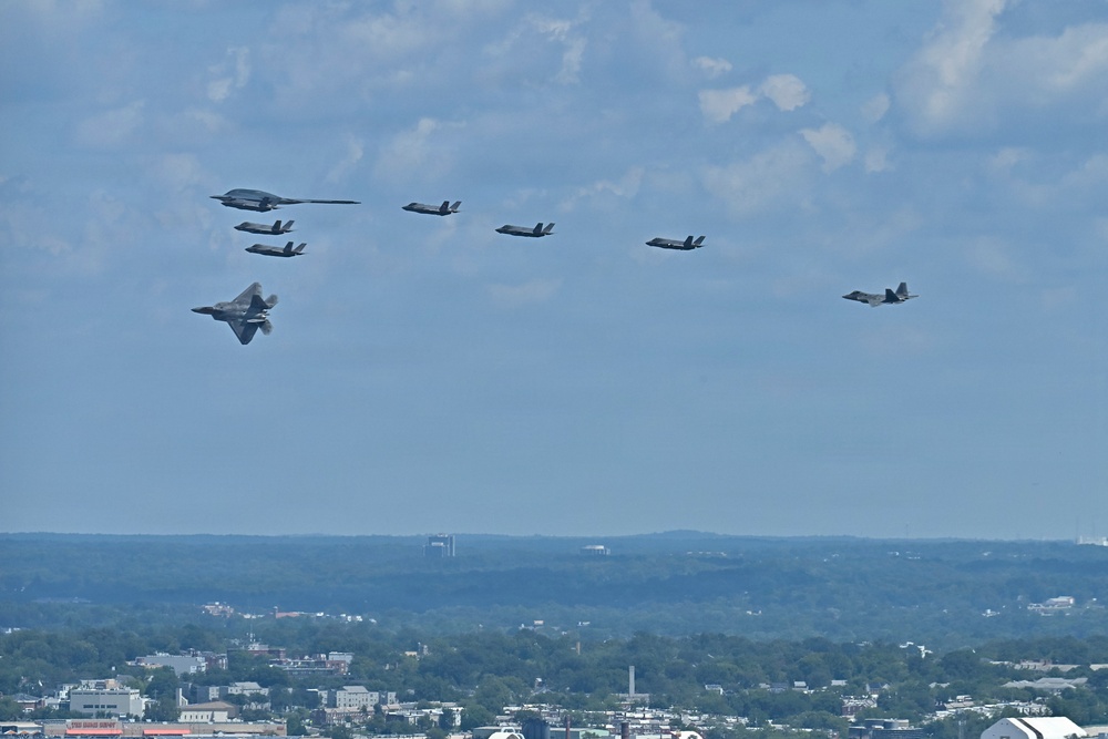 A formation of multiple U.S. military stealth aircraft flying over an urban/suburban landscape under a partly cloudy sky. The formation appears to include a mix of aircraft types including what appears to be a B-2 Spirit stealth bomber, F-22 Raptors, and F-35 Lightning II fighters. This appears to be a ceremonial or demonstration flyover, likely over the Washington D.C. area based on the visible urban landscape.