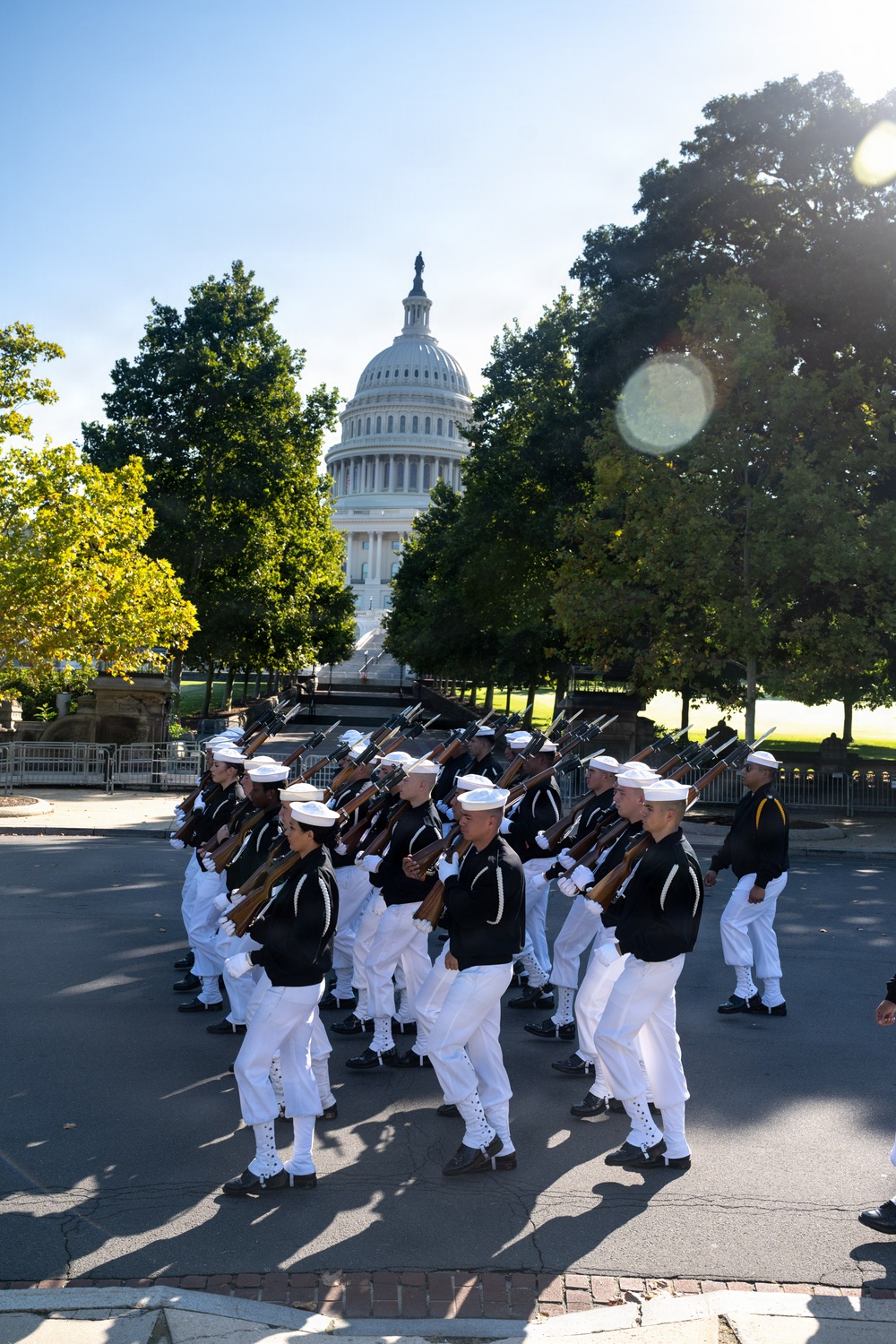 A military drill team in U.S. Navy dress uniforms marches in formation while carrying rifles in front of the United States Capitol building in Washington, D.C. The personnel are wearing distinctive black jumpers with white trousers and white sailor caps, consistent with the U.S. Navy Ceremonial Guard or a similar precision drill unit. The image was taken on a clear, sunny day with lens flare visible in the upper right corner.