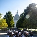 A military drill team in U.S. Navy dress uniforms marches in formation while carrying rifles in front of the United States Capitol building in Washington, D.C. The personnel are wearing distinctive black jumpers with white trousers and white sailor caps, consistent with the U.S. Navy Ceremonial Guard or a similar precision drill unit. The image was taken on a clear, sunny day with lens flare visible in the upper right corner.
