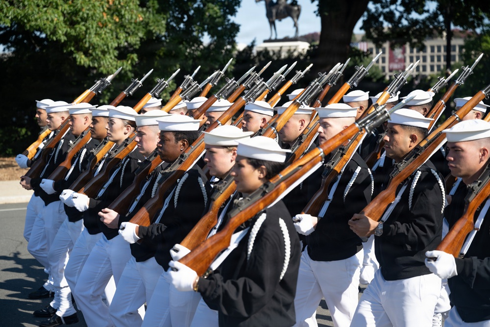A military ceremonial unit is marching in formation, carrying rifles with bayonets fixed. The personnel are dressed in formal Navy dress uniforms consisting of white caps, black jumpers, and white trousers with white gloves. A equestrian statue and buildings are visible in the background, suggesting this is a formal parade or ceremonial event in an urban setting.