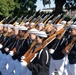 A military ceremonial unit is marching in formation, carrying rifles with bayonets fixed. The personnel are dressed in formal Navy dress uniforms consisting of white caps, black jumpers, and white trousers with white gloves. A equestrian statue and buildings are visible in the background, suggesting this is a formal parade or ceremonial event in an urban setting.