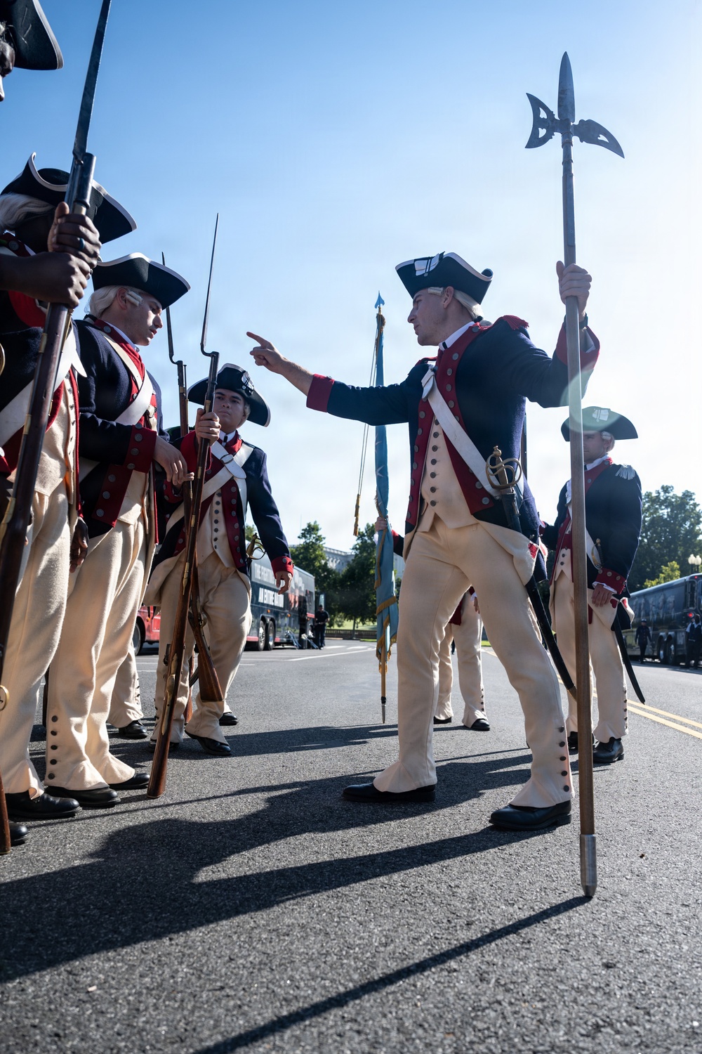A group of individuals dressed in 18th-century American Revolutionary War-era military uniforms are assembled on a paved road, likely during a parade or ceremonial event. One figure holding a halberd (pole weapon) appears to be giving instructions or directions to the formation. The setting appears to be Washington D.C., based on visible background architecture and landscape.