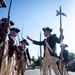 A group of individuals dressed in 18th-century American Revolutionary War-era military uniforms are assembled on a paved road, likely during a parade or ceremonial event. One figure holding a halberd (pole weapon) appears to be giving instructions or directions to the formation. The setting appears to be Washington D.C., based on visible background architecture and landscape.