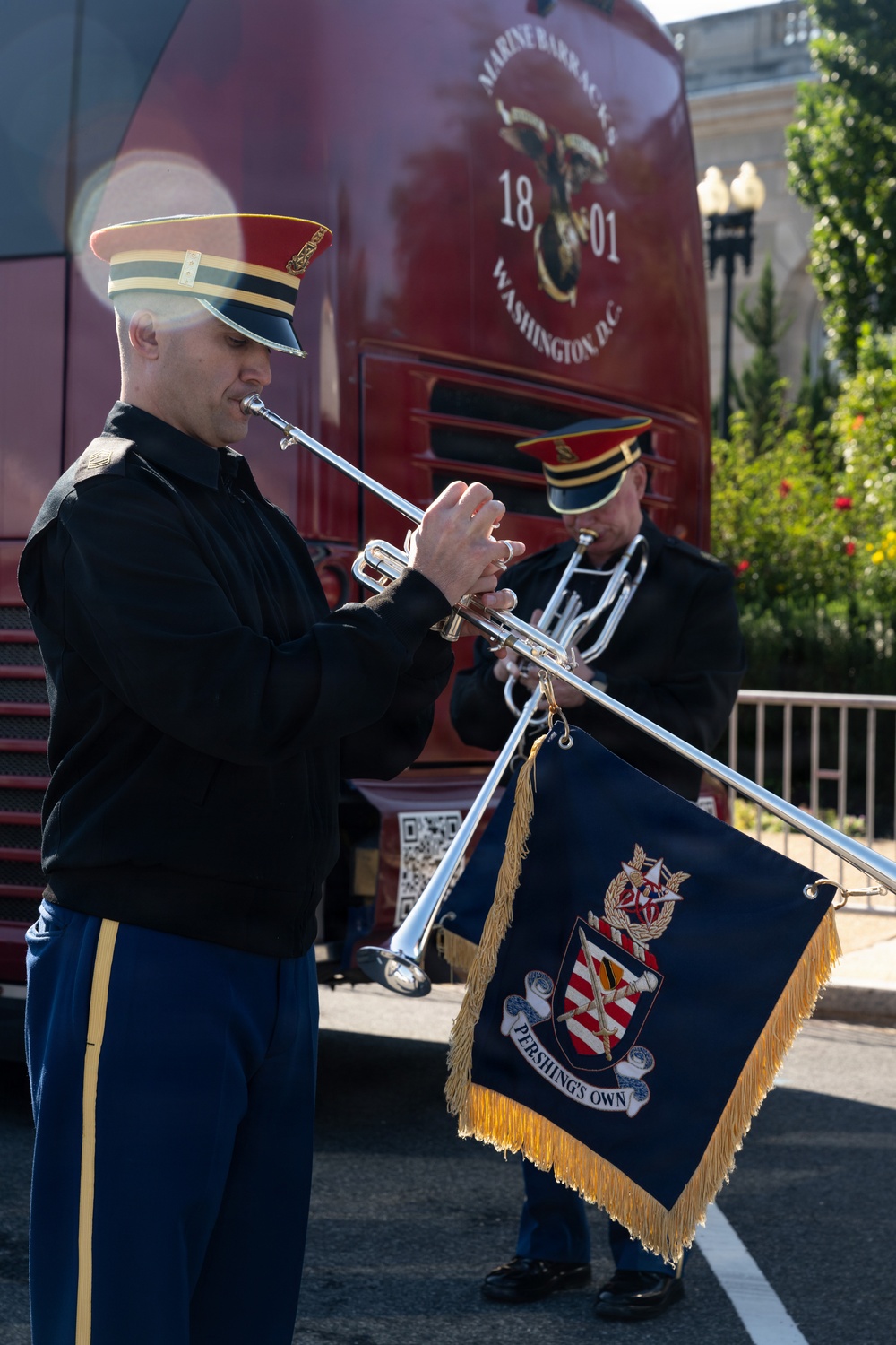 Two military musicians in dress uniforms are playing silver trumpets/herald trumpets outdoors. One trumpet bears a navy blue ceremonial banner with gold fringe reading 'Pershing's Own' with a coat of arms. They are positioned in front of a red bus or vehicle marked 'Marine Barracks Washington D.C. 1801.'