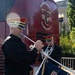 Two military musicians in dress uniforms are playing silver trumpets/herald trumpets outdoors. One trumpet bears a navy blue ceremonial banner with gold fringe reading 'Pershing's Own' with a coat of arms. They are positioned in front of a red bus or vehicle marked 'Marine Barracks Washington D.C. 1801.'