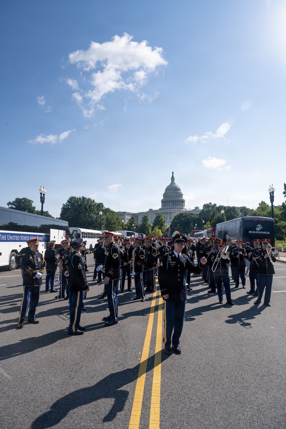 The United States Army Band is assembled on a street in Washington, D.C., with the U.S. Capitol Building prominently visible in the background. Band members are dressed in formal military dress uniforms with red-plumed caps, and a conductor stands at the front center on the double yellow center line. Support vehicles including a labeled 'The United States Army Band' bus and a Marines bus are parked along the sides.