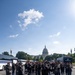 The United States Army Band is assembled on a street in Washington, D.C., with the U.S. Capitol Building prominently visible in the background. Band members are dressed in formal military dress uniforms with red-plumed caps, and a conductor stands at the front center on the double yellow center line. Support vehicles including a labeled 'The United States Army Band' bus and a Marines bus are parked along the sides.