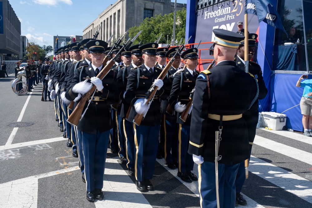 A U.S. Army ceremonial honor guard unit is performing a drill exhibition on a city street during what appears to be the 'Freedom 25 Grand Prix' event. Soldiers in formal dress uniforms are holding rifles with bayonets fixed, standing in formation while being inspected or commanded by an officer. The event appears to take place in Washington, D.C., based on the neoclassical architecture visible in the background.