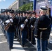 A U.S. Army ceremonial honor guard unit is performing a drill exhibition on a city street during what appears to be the 'Freedom 25 Grand Prix' event. Soldiers in formal dress uniforms are holding rifles with bayonets fixed, standing in formation while being inspected or commanded by an officer. The event appears to take place in Washington, D.C., based on the neoclassical architecture visible in the background.