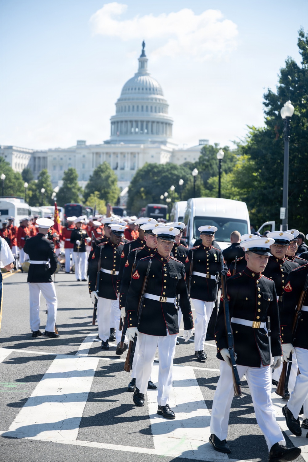 United States Marines in dress blues are marching in a formal parade or ceremonial procession on a street in Washington, D.C., with the U.S. Capitol Building prominently visible in the background. The Marines are in full dress uniform carrying rifles, accompanied by a band in red uniforms visible behind them. The scene depicts an official military ceremonial event on a clear, sunny day.