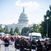 United States Marines in dress blues are marching in a formal parade or ceremonial procession on a street in Washington, D.C., with the U.S. Capitol Building prominently visible in the background. The Marines are in full dress uniform carrying rifles, accompanied by a band in red uniforms visible behind them. The scene depicts an official military ceremonial event on a clear, sunny day.