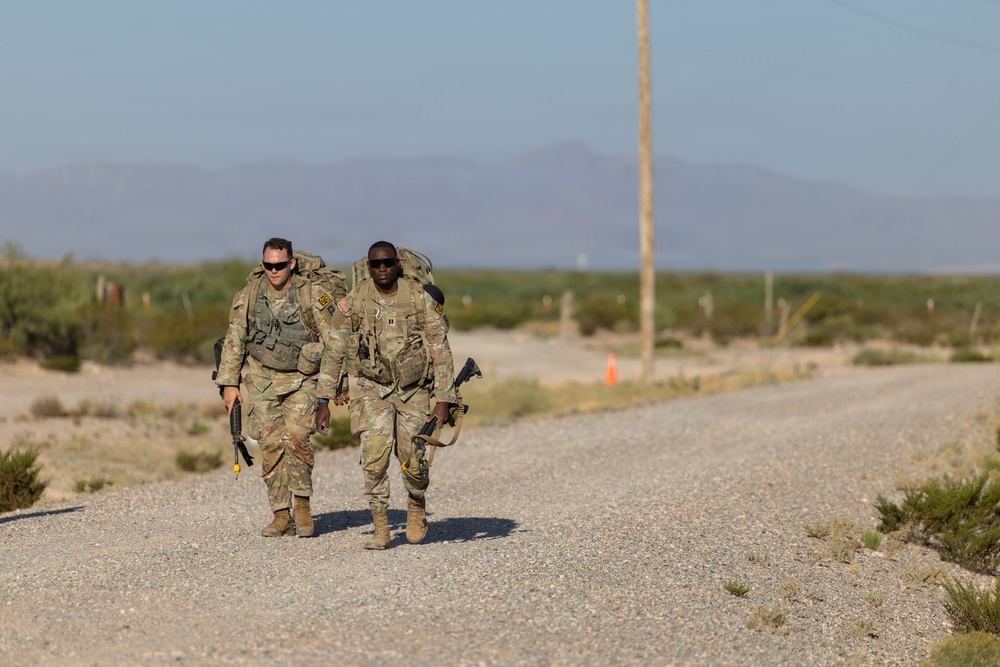 Two soldiers in US Army OCP (Operational Camouflage Pattern) uniforms are conducting a ruck march along a gravel road in an arid desert environment. Both are carrying large rucksacks and appear to be armed with rifles. The setting appears to be a military training area based on the terrain and road markings visible in the background.