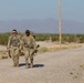 Two soldiers in US Army OCP (Operational Camouflage Pattern) uniforms are conducting a ruck march along a gravel road in an arid desert environment. Both are carrying large rucksacks and appear to be armed with rifles. The setting appears to be a military training area based on the terrain and road markings visible in the background.