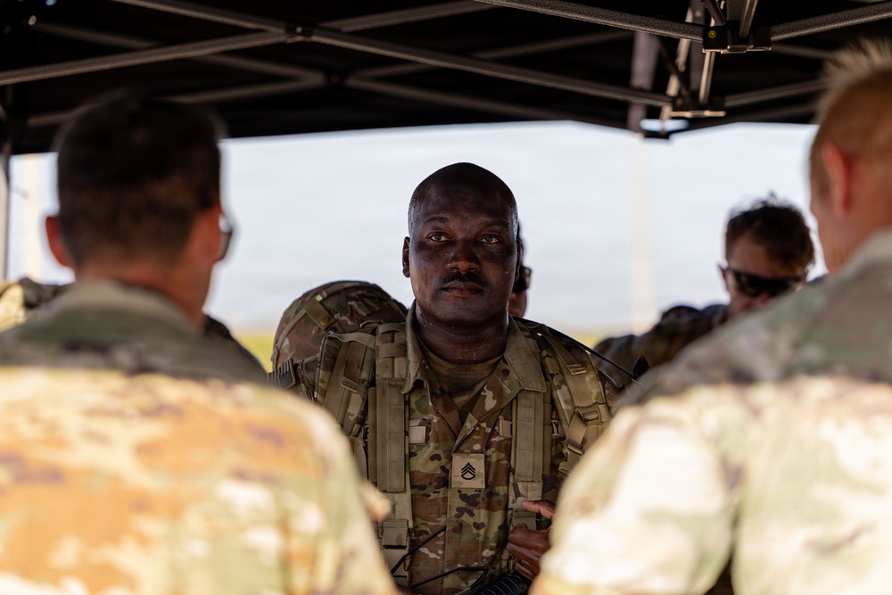 A U.S. Army Staff Sergeant in OCP (Operational Camouflage Pattern) uniform is addressing a small group of soldiers under a pop-up canopy shelter in an outdoor field environment. The central figure appears to be conducting a briefing or after-action review, facing the camera while speaking to personnel whose backs are turned. The setting suggests a field training exercise or operational environment.