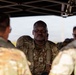 A U.S. Army Staff Sergeant in OCP (Operational Camouflage Pattern) uniform is addressing a small group of soldiers under a pop-up canopy shelter in an outdoor field environment. The central figure appears to be conducting a briefing or after-action review, facing the camera while speaking to personnel whose backs are turned. The setting suggests a field training exercise or operational environment.