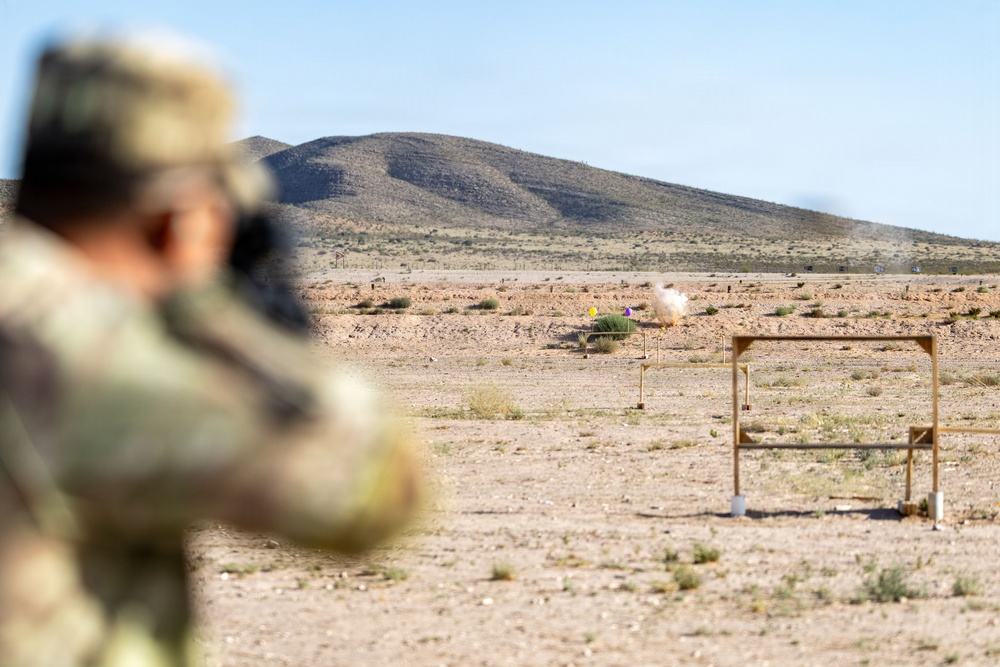 A military or law enforcement personnel in camouflage uniform is firing at a shooting range in an arid desert environment. A visible impact or explosion can be seen at a downrange target, likely an exploding reactive target. The scene takes place against a backdrop of desert terrain with a low hill in the distance.
