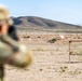 A military or law enforcement personnel in camouflage uniform is firing at a shooting range in an arid desert environment. A visible impact or explosion can be seen at a downrange target, likely an exploding reactive target. The scene takes place against a backdrop of desert terrain with a low hill in the distance.